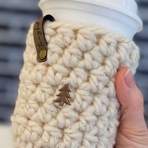 May include: White crocheted coffee cup cozy with a gold thread detail and a small brown tree charm. The cozy has a brown leather strap with the text "Hot Lava" on it.