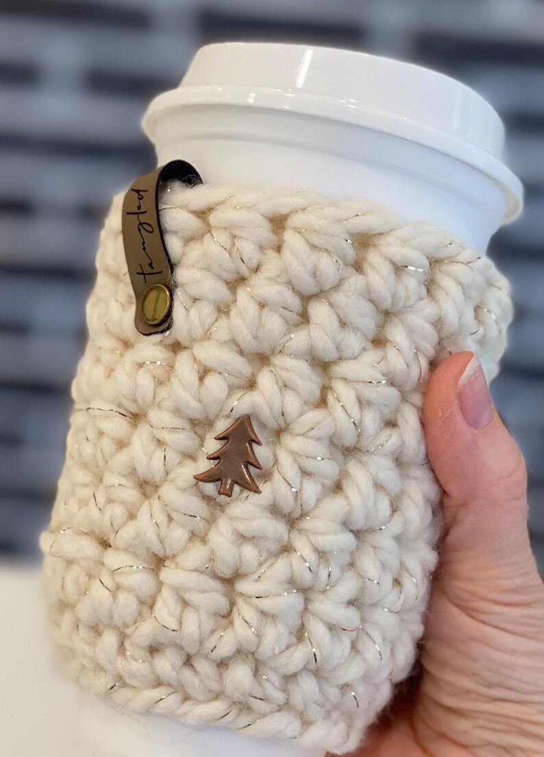 May include: White crocheted coffee cup cozy with a gold thread detail and a small brown tree charm. The cozy has a brown leather strap with the text "Hot Lava" on it.
