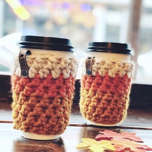May include: Two crocheted coffee cup cozies in orange, yellow, and white with a brown leather tag that says "TDBK". The cozies are sitting on a wooden table with fall leaves.