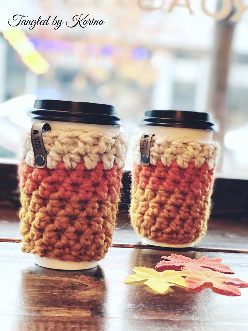 May include: Two crocheted coffee cup cozies in orange, yellow, and white with a brown leather tag that says "TDBK". The cozies are sitting on a wooden table with fall leaves.