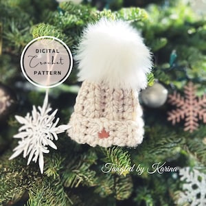 May include: A digital crochet pattern for a white beanie with a faux fur pom pom. The beanie has a brown leather maple leaf accent. The beanie is hanging on a Christmas tree.