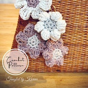 May include: Close-up of several handmade crochet flower ornaments in shades of white and mauve. The flowers are attached to a woven basket. The text "Crochet Pattern" and "Tangled by Karina" are visible in a circular design.