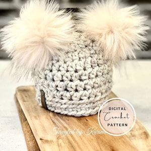 Pode incluir: Um gorro de crochê branco com dois pompons fofinhos. O gorro está sobre uma superfície de madeira com o texto "DIGITAL Crochet PATTERN" e "Tangled by Karina" visível.
