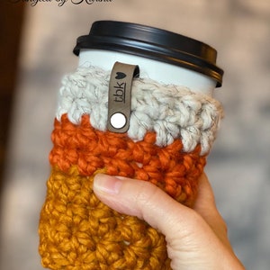 May include: A crocheted coffee cup cozy in orange, white, and brown with a leather strap that says "tbk" with a heart.