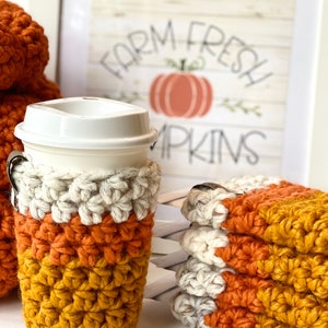 May include: A set of three crocheted coffee cup cozies in orange, white, and mustard yellow. The cozies are stacked on top of each other. The cozies are in front of a framed print that says "Farm Fresh Pumpkins".