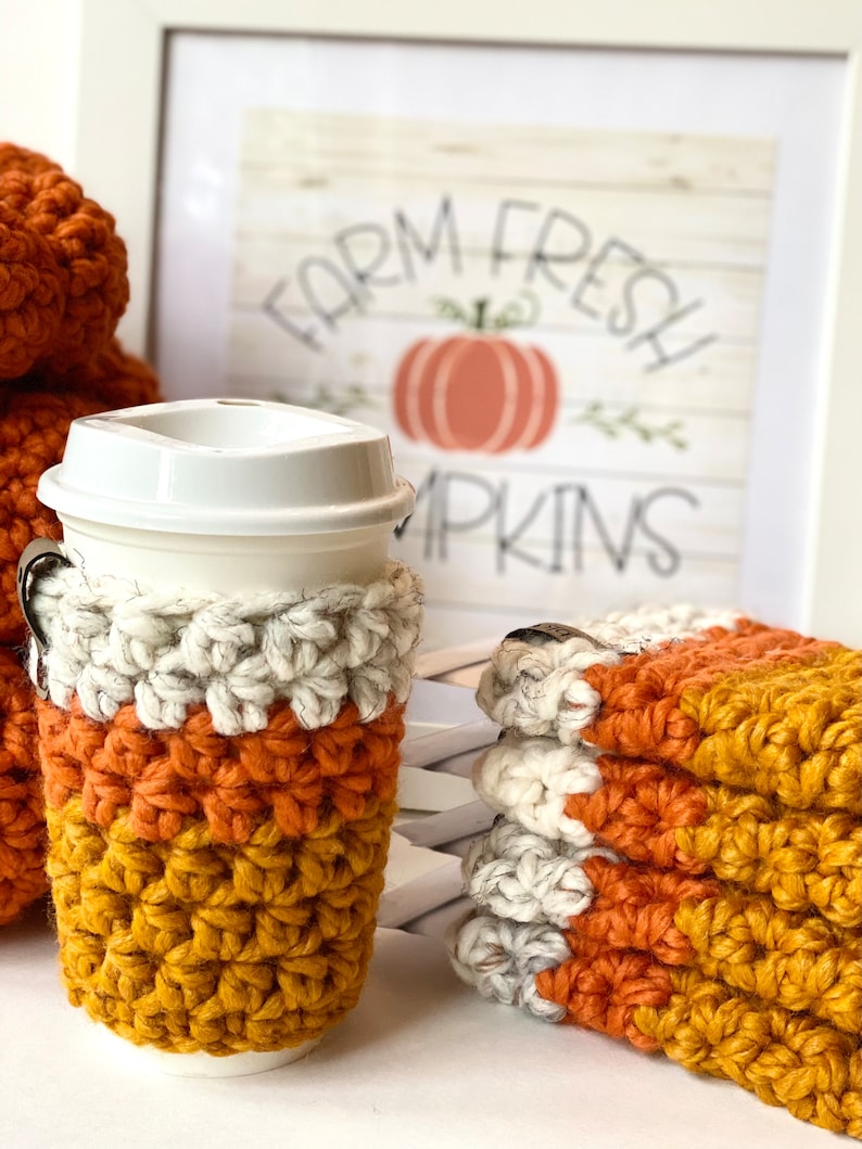 May include: A set of three crocheted coffee cup cozies in orange, white, and mustard yellow. The cozies are stacked on top of each other. The cozies are in front of a framed print that says "Farm Fresh Pumpkins".