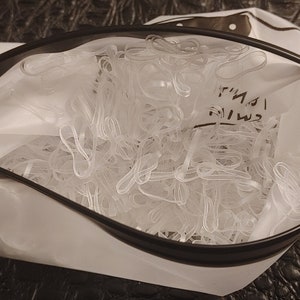 May include: A clear, zippered pouch filled with numerous clear hair ties. The pouch is white with a black zipper and trim. Text is visible on the pouch. The background is a dark, textured surface.
