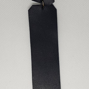 Könnte beinhalten: Ein schwarzer Leder-Lesezeichen mit geometrischem Design. Das Lesezeichen hat einen rechteckigen Körper mit einem kleinen, abgewinkelten Oberteil, das durch einen Metallring verbunden ist. Das Leder hat eine strukturierte Oberfläche.