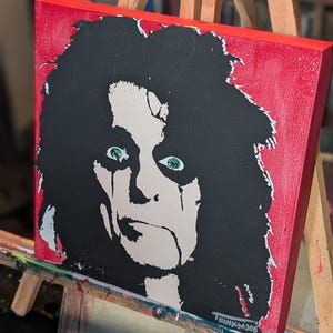 Puede incluir: Arte en lienzo cuadrado que presenta un retrato estilizado con cabello negro, ojos verdes y pintura facial blanca sobre un fondo rojo. La obra de arte se exhibe en un caballete de madera. La firma del artista, "THINK306", es visible.