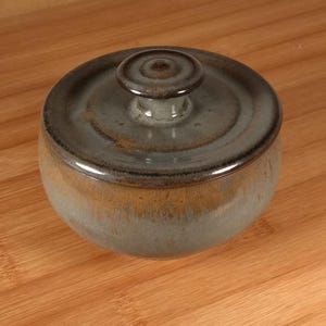 May include: A round ceramic lidded jar featuring a smooth, glossy finish with a gradient of muted gray and earthy brown tones. The lid includes a small, centered knob handle, and the overall design displays a consistent, glazed texture throughout.