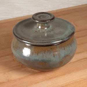 May include: A small, round ceramic jar with a lid. The jar is glazed in shades of brown, teal, and gray. The lid has a small, rounded handle. The jar is sitting on a wooden surface.