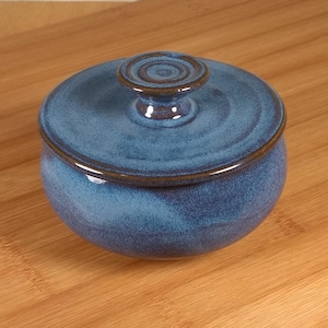 May include: A round ceramic lidded jar featuring a deep blue glaze with subtle variations in tone. The lid has a small, centered knob handle with concentric circular ridges. The base of the jar is rounded, and the rim is finished in a dark brown.