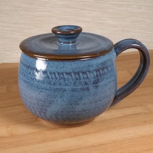 May include: A blue ceramic mug with a lid. The mug has a rounded shape with a handle and a lid with a small knob. The mug is glazed with a blue color and has a brown rim.