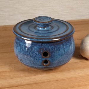May include: A round, blue ceramic garlic keeper with a matching lid and a small knob handle. The container features two circular ventilation holes on the front and a glossy, mottled blue glaze with darker brown edges around the rim and lid.