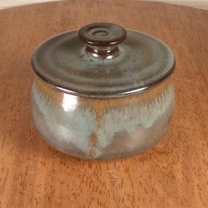 May include: A small, round ceramic jar with a matching lid featuring a glossy finish. The glaze displays a blend of earthy brown and muted blue-green tones that transition smoothly across the surface. The lid has a small, centred knob handle.
