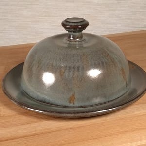 May include: A ceramic butter dish with a domed lid and a small knob handle. The dish and lid are a muted teal color with brown accents. The dish sits on a round base, perfect for serving butter.