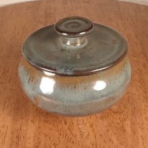 May include: A round ceramic lidded jar featuring a glossy glaze with blended tones of muted blue, grey, and earthy brown. The lid has a small, centered knob handle, and the body of the jar has a smooth, curved silhouette with a reflective surface.