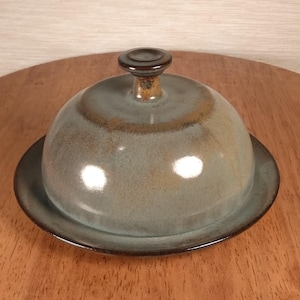 May include: A ceramic butter dish with a domed lid and a circular base. The dish is glazed in shades of blue and brown, with a dark brown knob on top. The dish is sitting on a wooden surface.