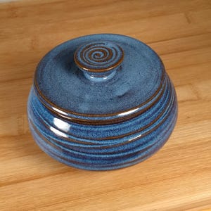 May include: A round ceramic lidded jar featuring a deep blue glaze with visible brown undertones along the horizontal ridges. The lid includes a small, circular knob with a spiral pattern etched into the center, matching the brown rim accents.