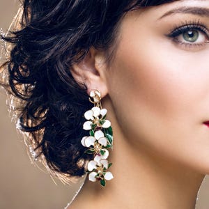 May include: A long drop earring featuring a series of white flower petals with green leaves and small gold-toned accents. The floral design is arranged in a vertical cluster, suspended from a small gold-toned circular stud at the top of the earlobe.