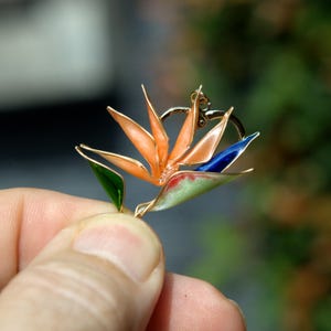 K&ouml;nnte beinhalten: Ein goldfarbener Metallanh&auml;nger in Form einer Paradiesvogelblume mit durchscheinenden orangefarbenen Bl&uuml;tenbl&auml;ttern, einem tiefblauen Akzent und einem hellgr&uuml;nen Blatt, alles umrandet von d&uuml;nnem Golddraht mit einer kleinen &Ouml;se an der Oberseite.