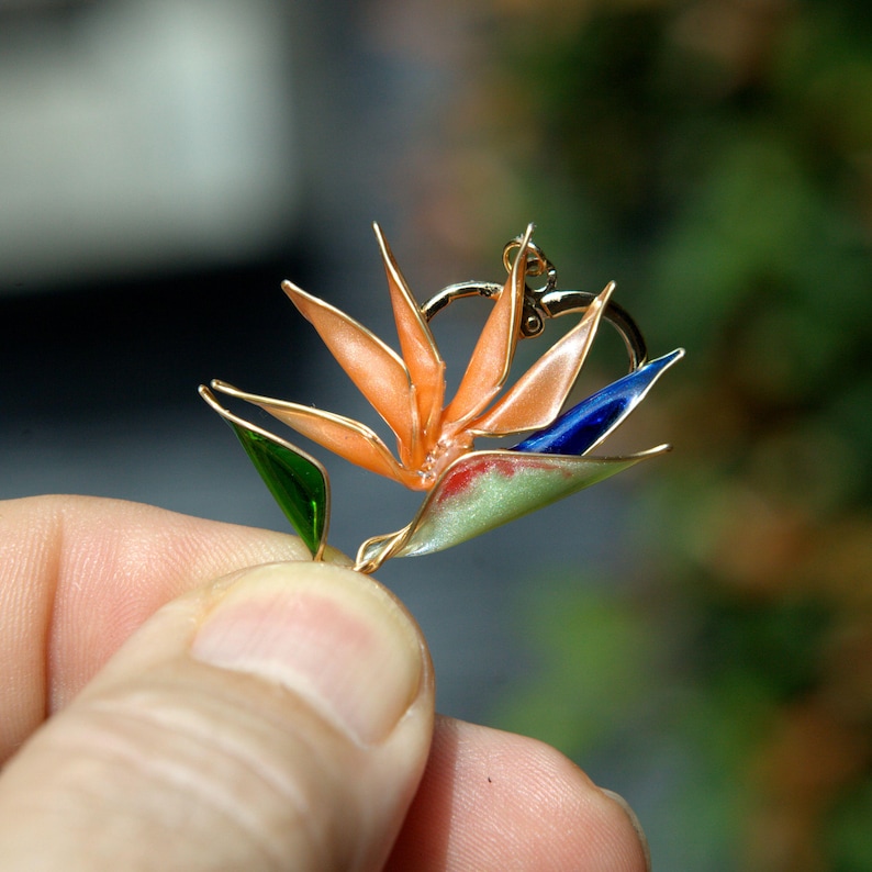 K&ouml;nnte beinhalten: Ein goldfarbener Metallanh&auml;nger in Form einer Paradiesvogelblume mit durchscheinenden orangefarbenen Bl&uuml;tenbl&auml;ttern, einem tiefblauen Akzent und einem hellgr&uuml;nen Blatt, alles umrandet von d&uuml;nnem Golddraht mit einer kleinen &Ouml;se an der Oberseite.