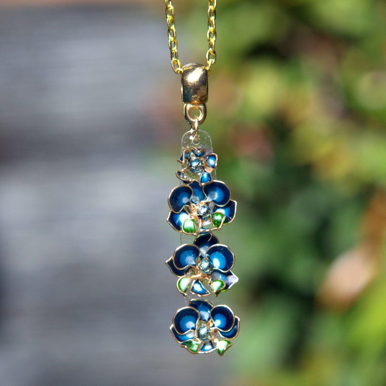 May include: A gold-toned necklace with a pendant featuring three blue flower charms with green and white accents. The flowers are enameled with a dark blue hue and have a gold-colored outline. The necklace is hanging against a blurred green background.