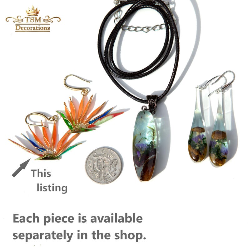 K&ouml;nnte beinhalten: Ein Schmuckset bestehend aus zwei Ohrringen in Form einer Paradiesvogelblume, einer langen ovalen Anh&auml;ngerkette mit eingebetteten botanischen Elementen und zwei tropfenf&ouml;rmigen Ohrringen mit Pilzen und violetten Blumen, neben einer 10-Cent-M&uuml;nze.