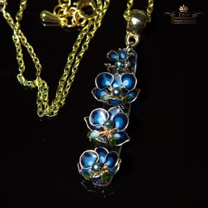 May include: A gold-colored chain necklace with a pendant featuring three blue flower charms. The flowers have dark blue petals with gold trim and a green center. The necklace is on a black surface.
