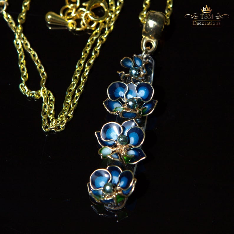 May include: A gold-colored chain necklace with a pendant featuring three blue flower charms. The flowers have dark blue petals with gold trim and a green center. The necklace is on a black surface.