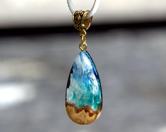 May include: A teardrop-shaped pendant featuring a combination of natural light-colored wood at the base and translucent blue and white resin at the top. The pendant is suspended from a white cord by a decorative gold-toned metal bail.