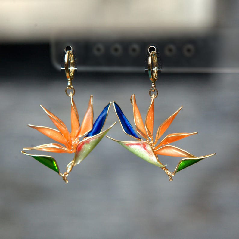 K&ouml;nnte beinhalten: Ein Paar Ohrh&auml;nger in Form von Paradiesvogelblumen. Jeder Ohrring besteht aus orangefarbenen, blauen und gr&uuml;nen Emaille-Bl&uuml;tenbl&auml;ttern mit goldfarbenen Metallr&auml;ndern. Die Ohrringe h&auml;ngen an kleinen, aufklappbaren goldfarbenen Creolen und fangen die leuchtenden Farben sowie die markante, spitze Form der tropischen Blume in einem detaillierten Design ein.
