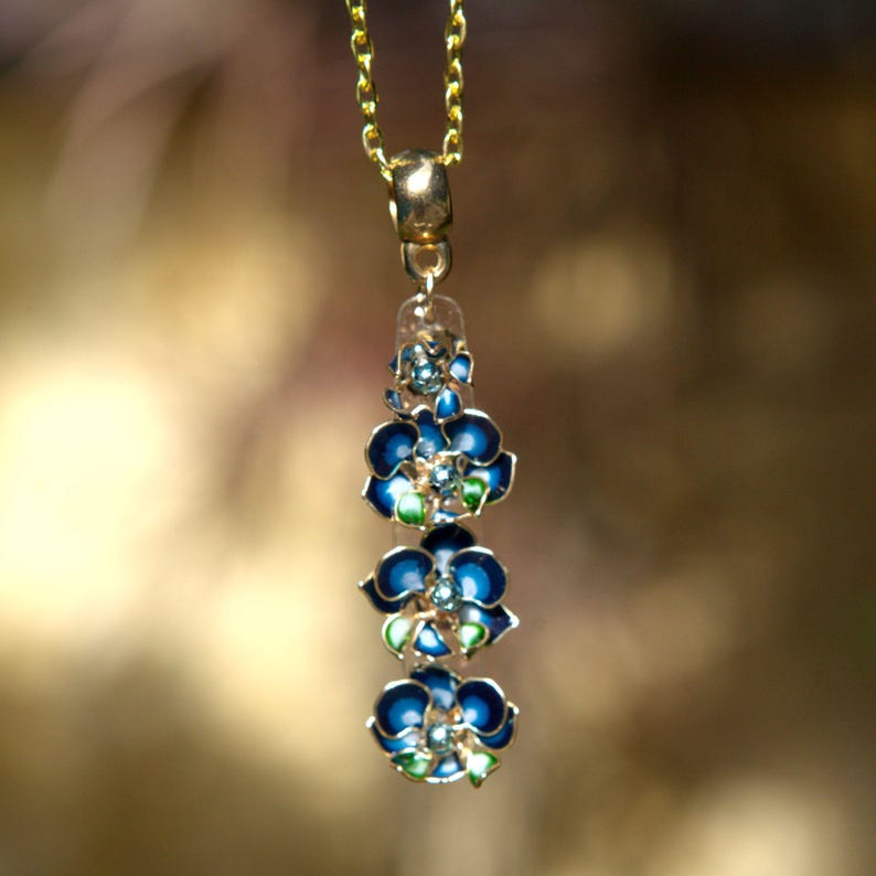 May include: A gold-toned necklace with a pendant featuring three blue flower charms with green and white accents. The flowers are enameled and have a detailed design. The chain is delicate and the background is blurred.