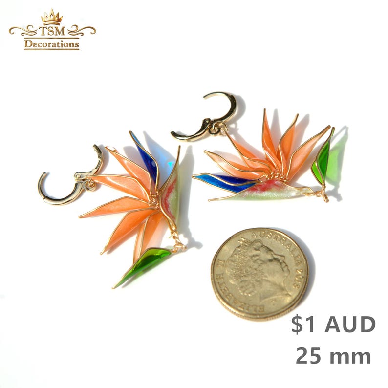 K&ouml;nnte beinhalten: Ein Paar goldfarbene Creolen in Form von Paradiesvogelblumen mit orangefarbenen, blauen und gr&uuml;nen Emaille-Details. Die Ohrringe sind neben einer 25 mm M&uuml;nze zur Gr&ouml;&szlig;enbestimmung abgebildet, wobei das TSM Decorations Logo zu sehen ist.