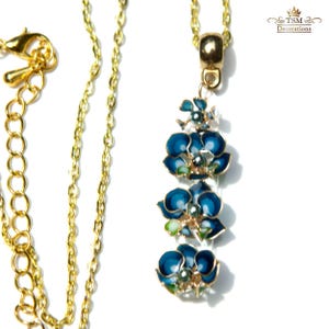 May include: A gold-toned necklace featuring a pendant with three blue flower designs. The flowers have dark blue petals with white and green accents. The necklace has a delicate chain and a clasp.
