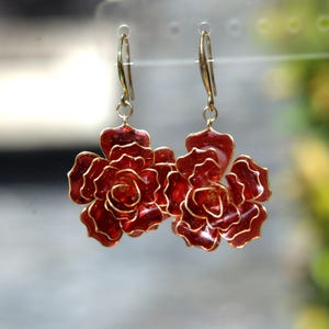 以下が含まれることがあります： ゴールドトーンのワイヤーエッジが付いた、深い赤色の半透明の花の形をしたチャームが特徴のドロップイヤリング。イヤリングはゴールドトーンの金属製フックから吊り下げられており、光を捉える重なり合った花びらのデザインが施されています。