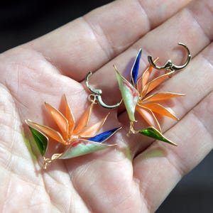 K&ouml;nnte beinhalten: Ein Paar goldfarbene Creolen mit Emaille-Anh&auml;ngern in Form einer Paradiesvogelblume. Das florale Design zeigt leuchtend orangefarbene Bl&uuml;tenbl&auml;tter, einen tiefblauen Akzent und eine blassgr&uuml;ne Basis mit roten Details an kleinen Klappb&uuml;geln.