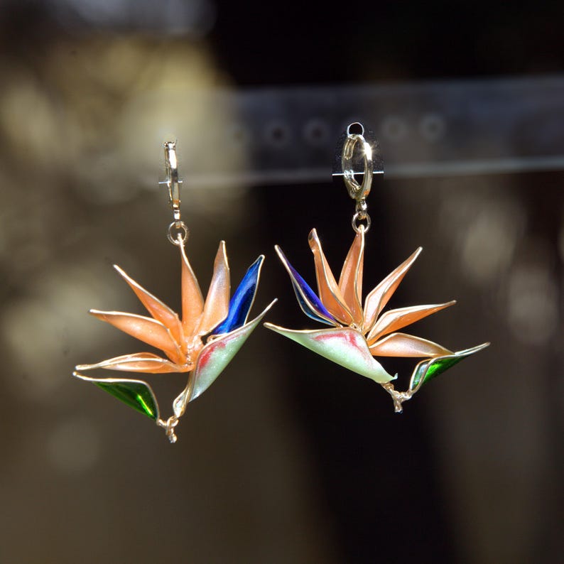 K&ouml;nnte beinhalten: Ein Paar goldfarbene Creolen mit h&auml;ngenden Anh&auml;ngern in Form von Paradiesvogelblumen. Die Blumenanh&auml;nger sind in leuchtenden Orange-, Blau- und Gr&uuml;nt&ouml;nen emailliert, wobei feine metallische Umrandungen die Bl&uuml;tenbl&auml;tter deutlich hervorheben.