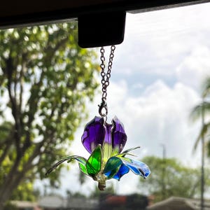 May include: A hanging ornament shaped like a flower with translucent purple, green, and blue petals. The flower is attached to a silver-toned metal chain with a lobster claw clasp, allowing it to be suspended from a rearview mirror or hook.