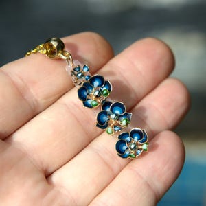 May include: A gold-toned necklace pendant featuring three blue flower charms with green and gold accents. The pendant is attached to a gold chain and is held in a hand.