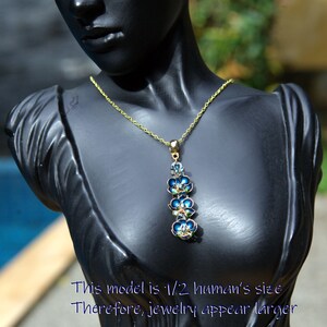 May include: A gold-toned necklace with a pendant featuring blue and green floral designs. The pendant is made of enamel and has a delicate, detailed appearance. The necklace is displayed on a black mannequin.