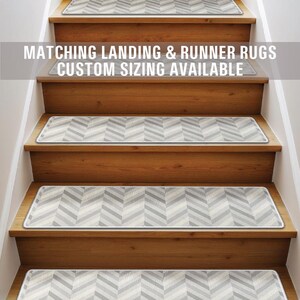Puede incluir: Un conjunto de peldaños de escalera con patrón de chevron gris y blanco con un borde blanco. El texto "Matching Landing & Runner Rugs Custom Sizing Available" se muestra sobre las escaleras.