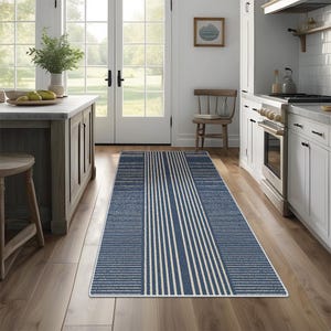 Puede incluir: Una alfombra de cocina rectangular con diseño de rayas azules y blancas. La alfombra presenta una sección central de rayas blancas verticales sobre un fondo azul, flanqueada por bordes a rayas azules y blancas texturizadas. Está colocada sobre un suelo de madera.