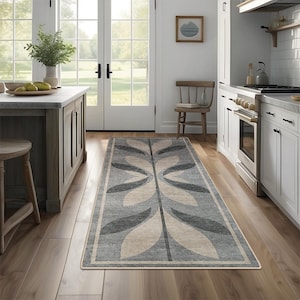 Puede incluir: Una alfombra rectangular con un diseño simétrico de hojas en tonos grises y beige. La alfombra está colocada sobre un suelo de madera en una cocina, con un borde blanco. La paleta de colores es neutra.