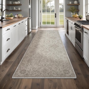 Puede incluir: Una alfombra de pasillo larga y rectangular con un intrincado patrón floral en tonos beige claro y gris, con un borde decorativo. La alfombra está colocada sobre un suelo de madera oscura en una cocina con armarios blancos y encimeras de madera.