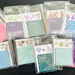 May include: Assortment of clear plastic bags containing various craft supplies, including cutting mats, stickers, and pens. The cutting mats are green, pink, and purple, and feature the word "Junesix" printed on them. The stickers feature various designs, including flowers, animals, and geometric patterns. The pens are black, purple, and pink.