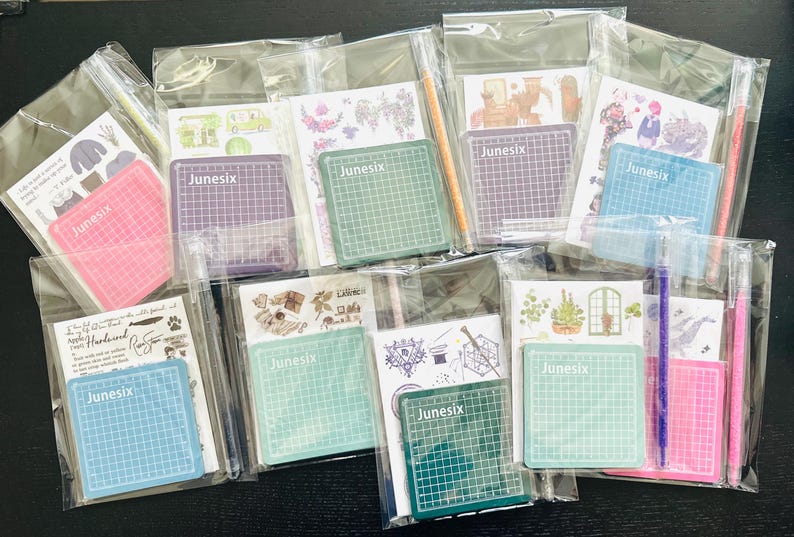 May include: Assortment of clear plastic bags containing various craft supplies, including cutting mats, stickers, and pens. The cutting mats are green, pink, and purple, and feature the word "Junesix" printed on them. The stickers feature various designs, including flowers, animals, and geometric patterns. The pens are black, purple, and pink.