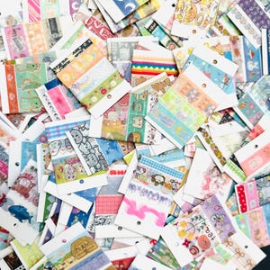 May include: A collection of decorative washi tape rolls in various colors and patterns. Designs include moons, bears, rainbows, and floral motifs. Each roll is mounted on a small card with a hole for hanging.