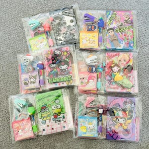 Op de afbeelding: Assortiment van kleurrijke kantoorartikelen sets met verschillende cartoonfiguren, waaronder Hello Kitty, Kuromi, Kerokerokeroppi en Disney-prinsessen. Elke set bevat een klein notitieboekje, een pen, een nietmachine en een sleutelhanger.