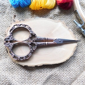 May include: Antique-style, ornate, copper-toned scissors with a floral design. The scissors have a silver blade and are resting on a wooden surface.